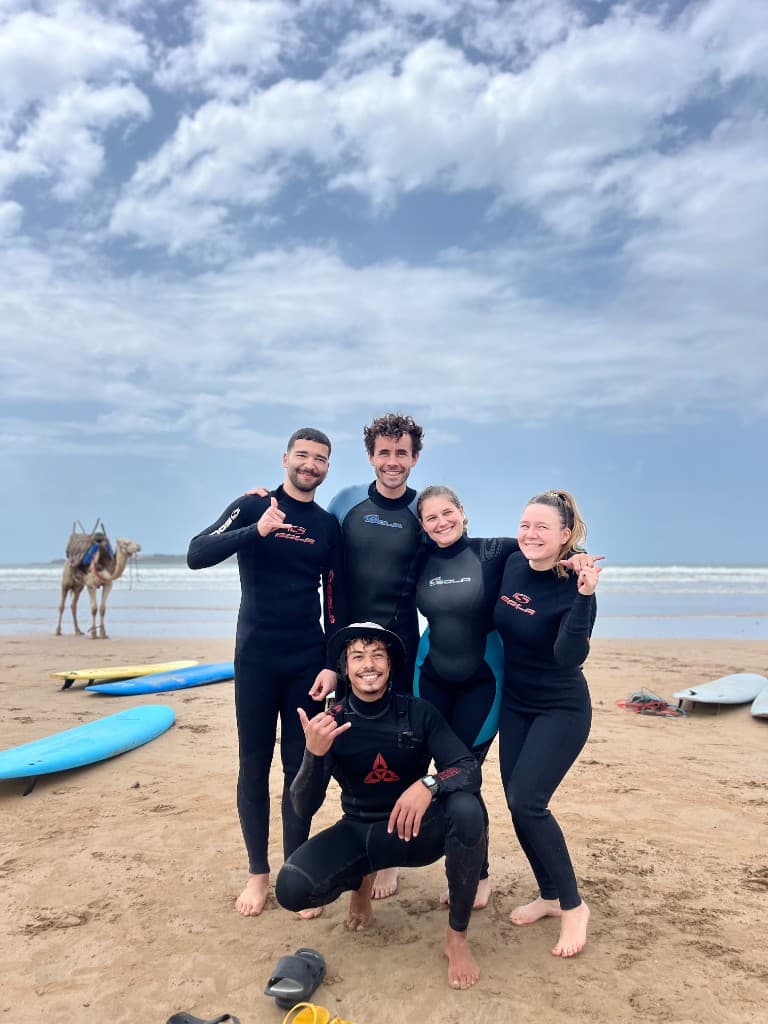 Five surf coaches and students in wetsuits on Essaouira beach with longboards on the sand, a camel, and small Atlantic waves under a bright sky — Morocco surf school team.