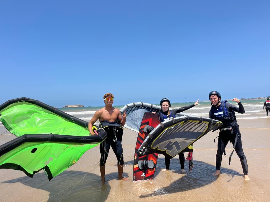 Kitesurf instructor and two students in helmets on Essaouira beach — neon green kite, wing, and board in shallow water, Morocco.