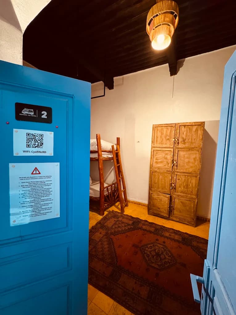 Surf camp dorm Essaouira view through blue wooden door log bunk beds Moroccan rug wooden lockers woven pendant lamp