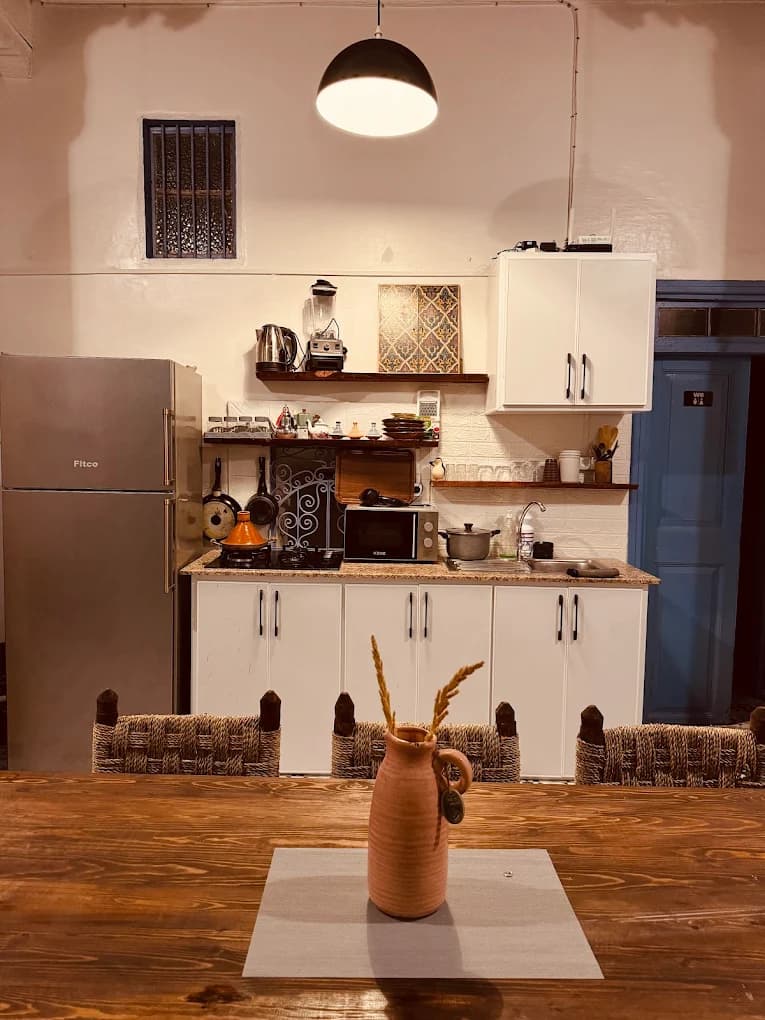 Surf camp Essaouira dining kitchen dark wood table woven straw chairs white cabinets stainless fridge tagine on stove microwave floating shelves pendant lamp