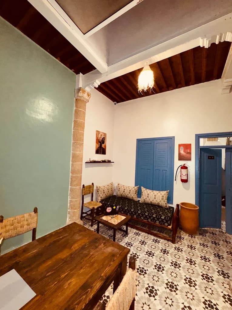 Cozy surf camp common room in Essaouira with Moroccan patterned floor tiles, blue wooden shutters, beamed ceiling, and rustic wooden seating
