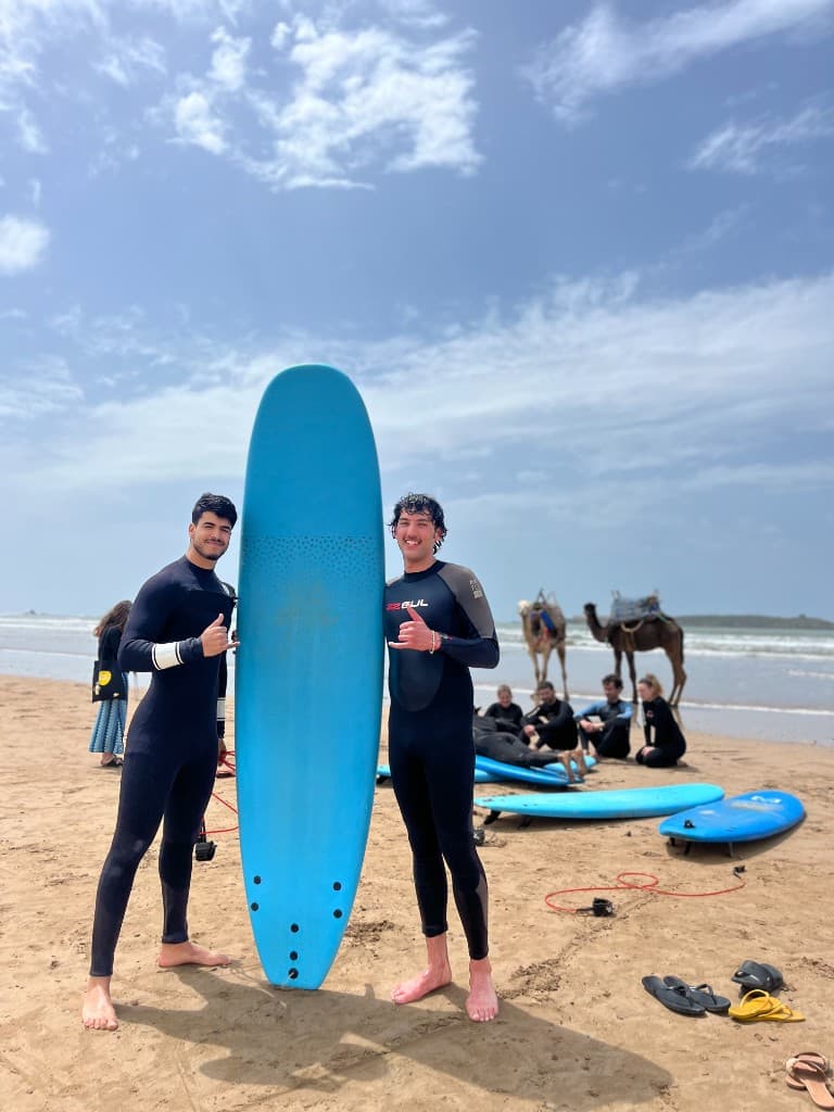 Two surf students in wetsuits with a blue longboard on Essaouira beach — camels and Atlantic swell in the background, Morocco.