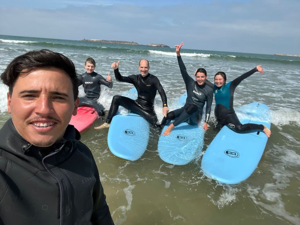 Surf instructor selfie with students on boards in shallow water at Essaouira — foamies, wetsuits, and smiles in the surf zone